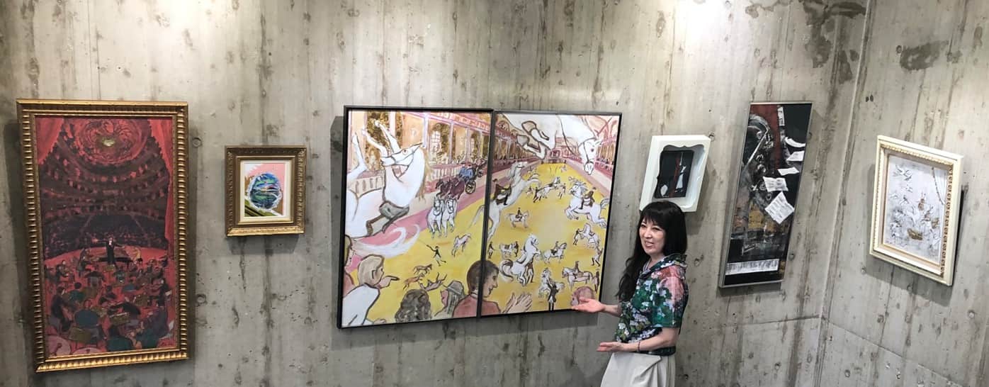 Une femme sourit en montrant deux grands tableaux représentant des chevaux et des personnes. Plusieurs autres œuvres d'art encadrées sont accrochées sur un mur en béton brut derrière elle.